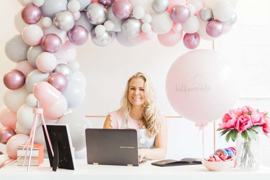 Balloonista branding shoot. Aurelija is smiling and sat at a white desk typing on her grey laptop. The desk has a pink lamp on the left, picture frame, notebook and pink flowers on the right. There is a giant pink 3ft branded balloon with the Balloonista logo in grey on the right side of her. A silver, grey, chrome pink, organic voluminous responsibly sourced latex balloon demi arch is in the background built from the left side of the image.