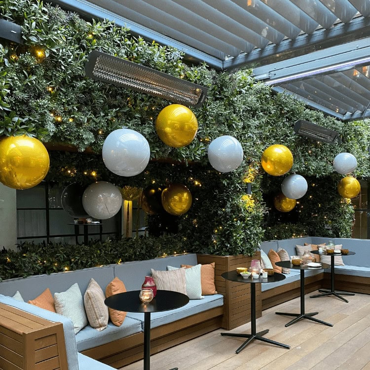 Gold and white balloons nestled among lush green foliage in an outdoor lounge area.
