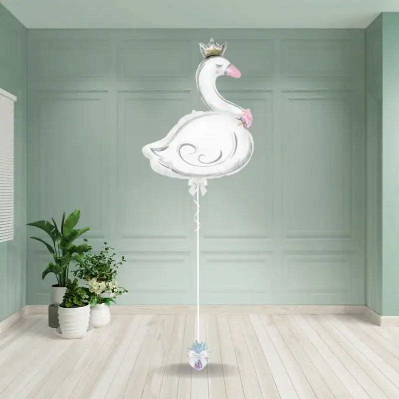 White swan-shaped balloon wearing a silver crown, with a pink beak and flower detail, attached to a weight with a white ribbon and bow.